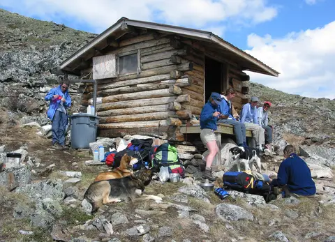 Kayalık yamaçtaki ahşap barınağın önünde dinlenen yürüyüşçüler ve yanlarında bir köpek