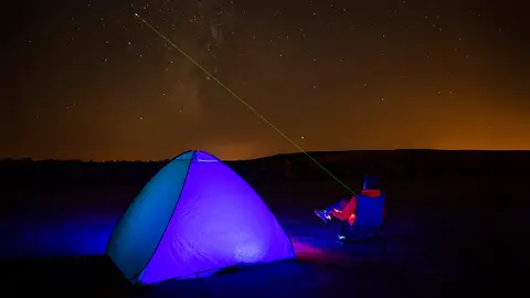 Yıldızlı gece gökyüzü altında içeriden mavi-mor ışıkla aydınlanan kubbe çadır ve yanında oturan kişi