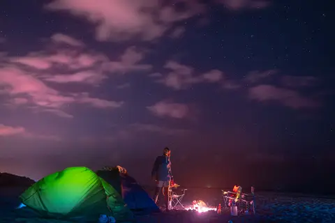 Bulutlu gece gökyüzü altında yeşil çadır, önünde küçük kamp ateşi ve katlanır sandalyeler